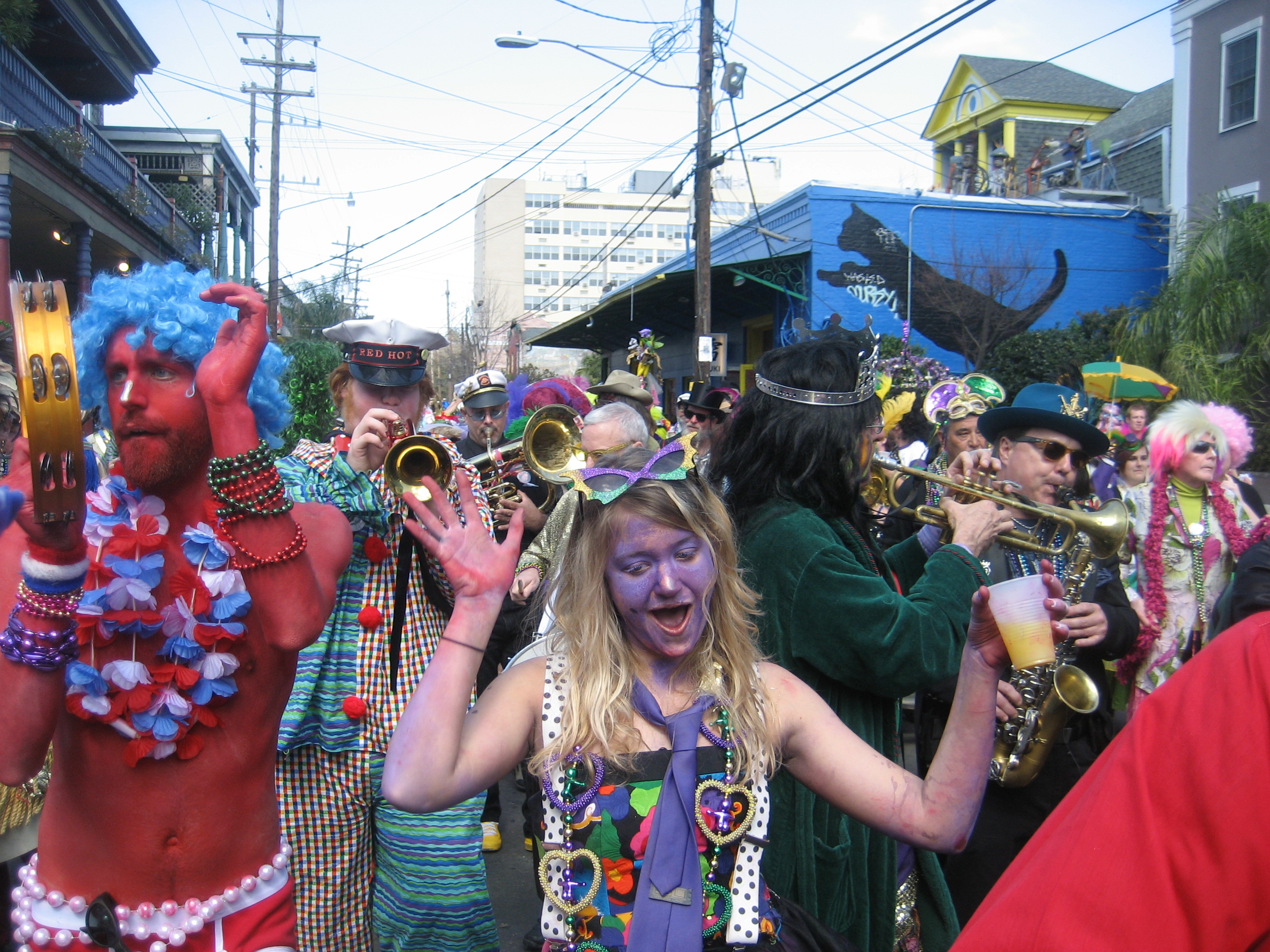 A image of a chaotic Mardi Gras street. Everyone is painted in colorful facepaint and dancing or having fun. In the front is a woman in purple face paint yelling. 